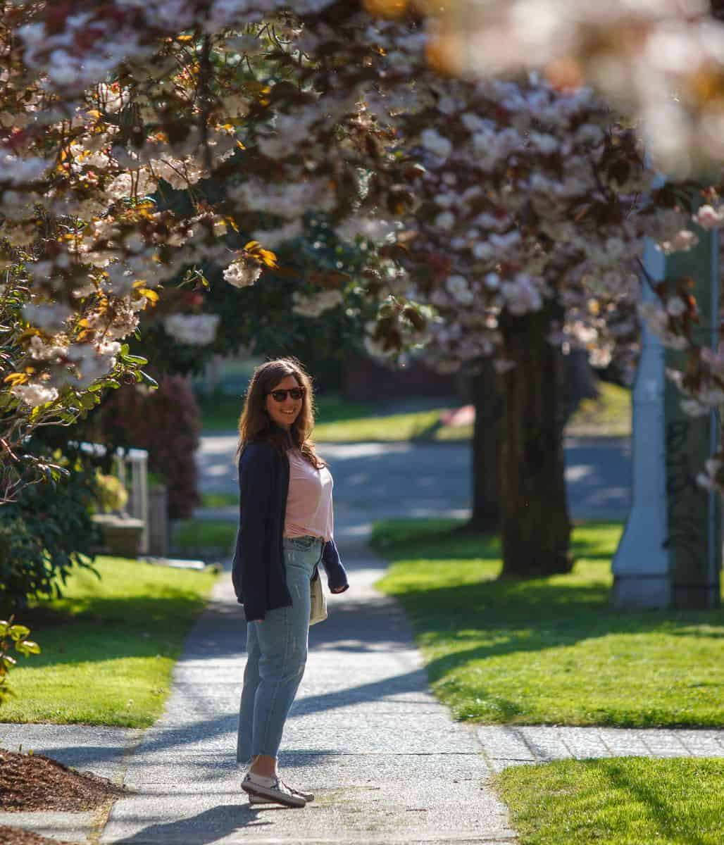 woman standing on a sidewalk under a canopy of light pink cherry blossoms in Victoria, BC