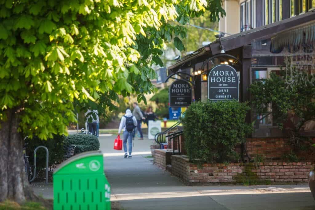 a pedestrian carrying a red shopping bag passes by a coffee shop patio that reads moka house on the awning